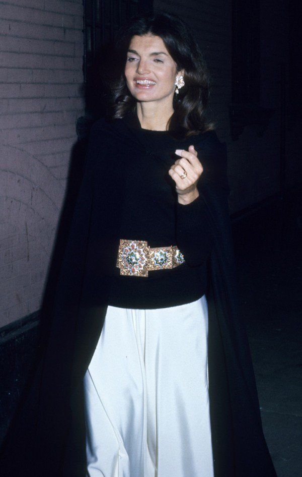 a woman standing in front of a brick wall wearing a black coat and white skirt