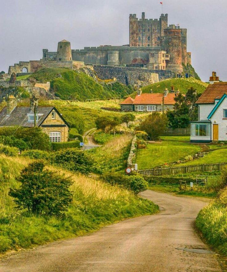 a dirt road leading to houses in front of a castle on top of a hill