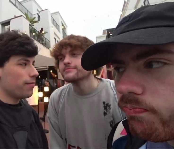 three young men standing next to each other in front of some buildings and one is looking at the camera