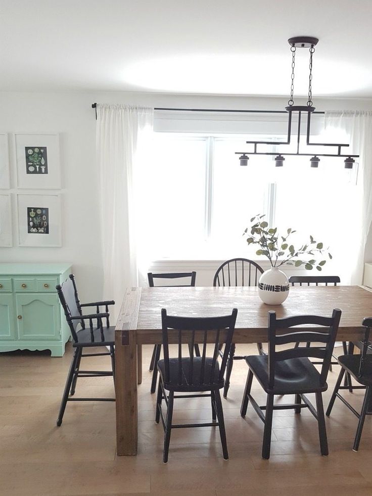 a dining room table with black chairs in front of a window and white curtains on the windowsill