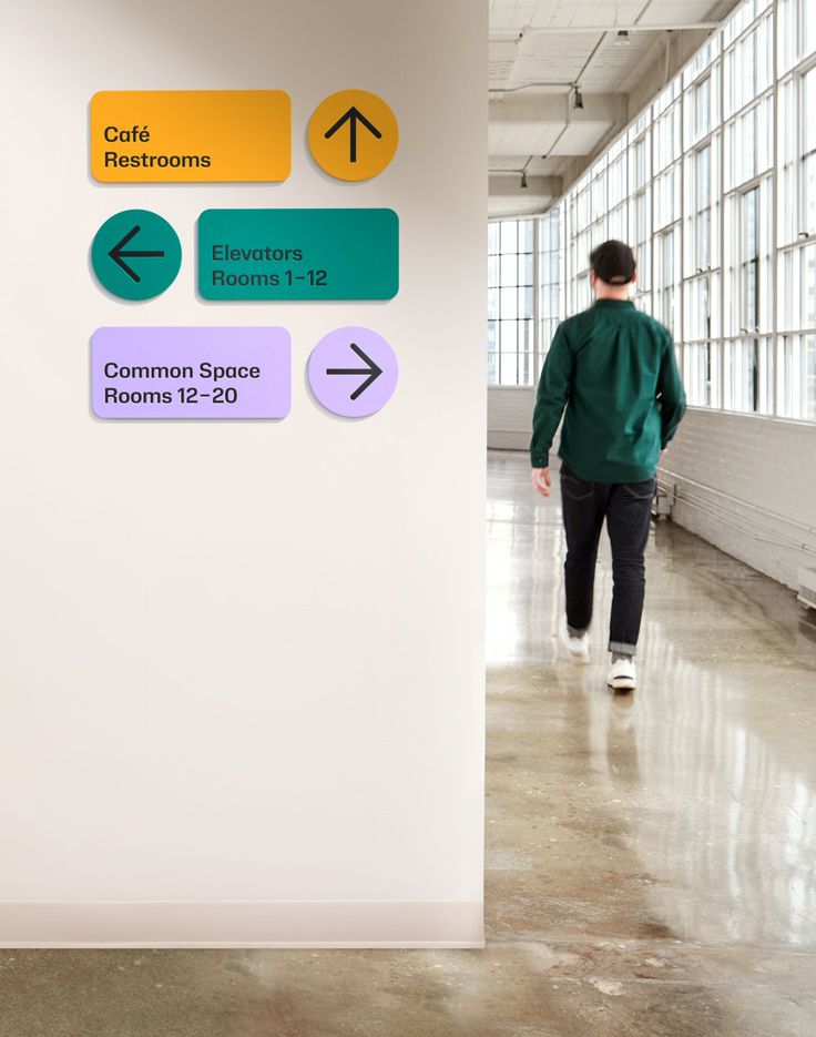 a man walking down a hallway next to a white wall with green and yellow signs on it