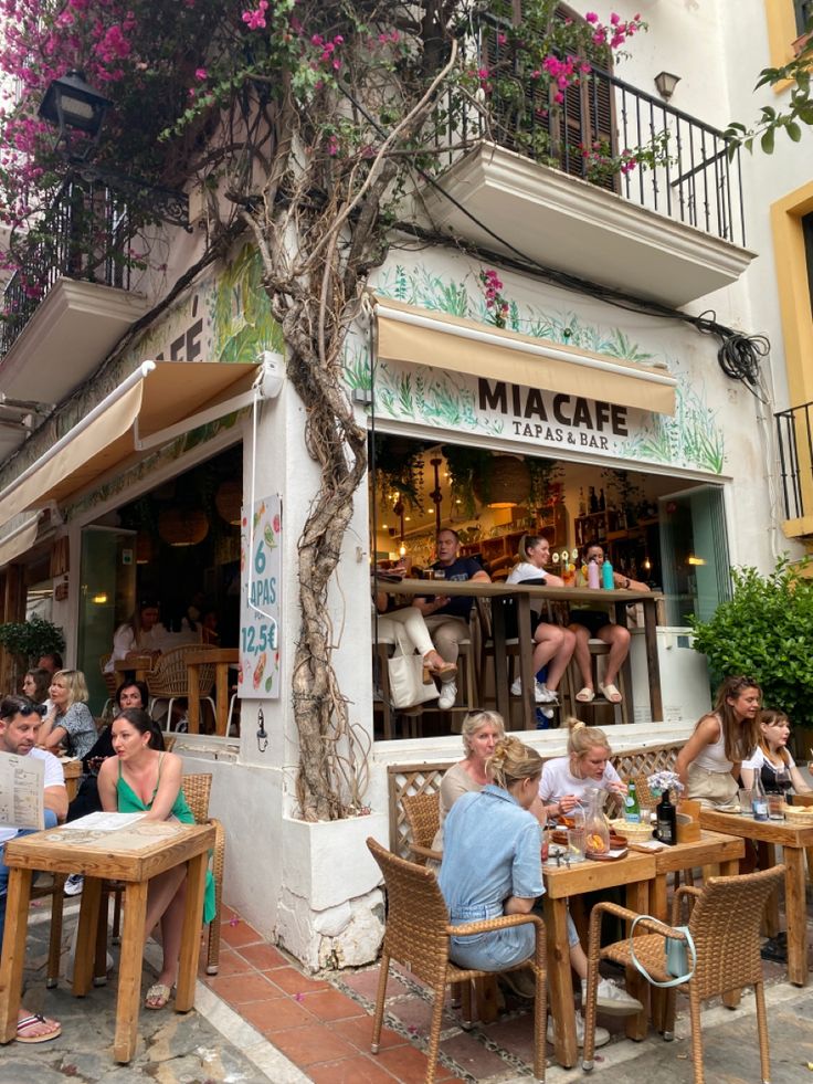 people are sitting at tables outside in front of a cafe with pink flowers on the tree