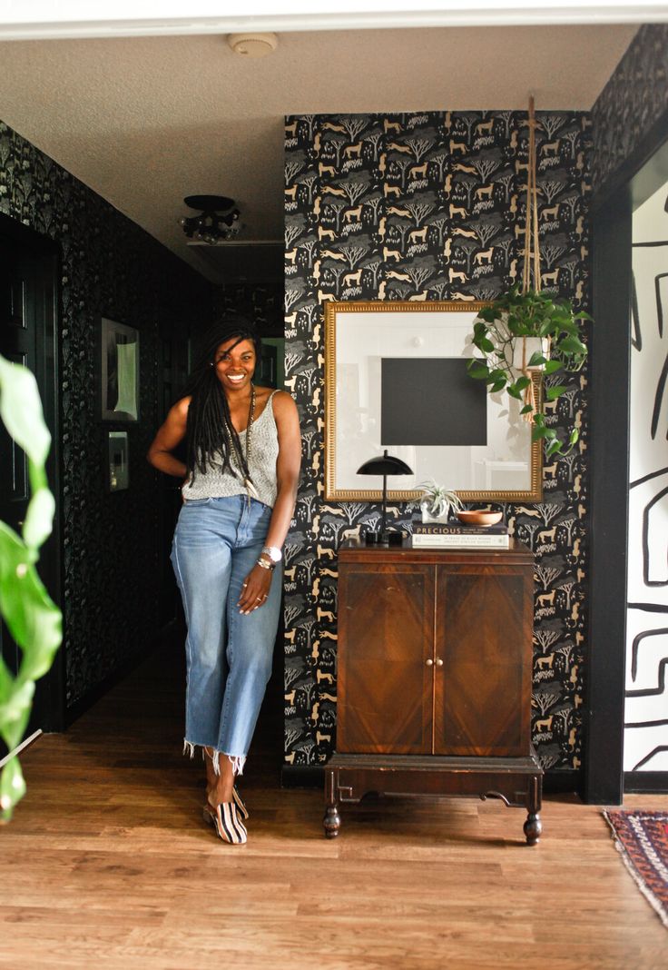 a woman standing next to a wooden cabinet in front of a wallpapered room