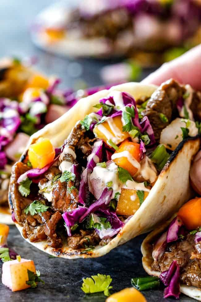 a hand holding a taco filled with meat and veggies next to other food