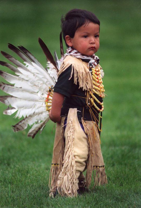 a little boy dressed in native american clothing with feathers on his head and chest, standing in the grass