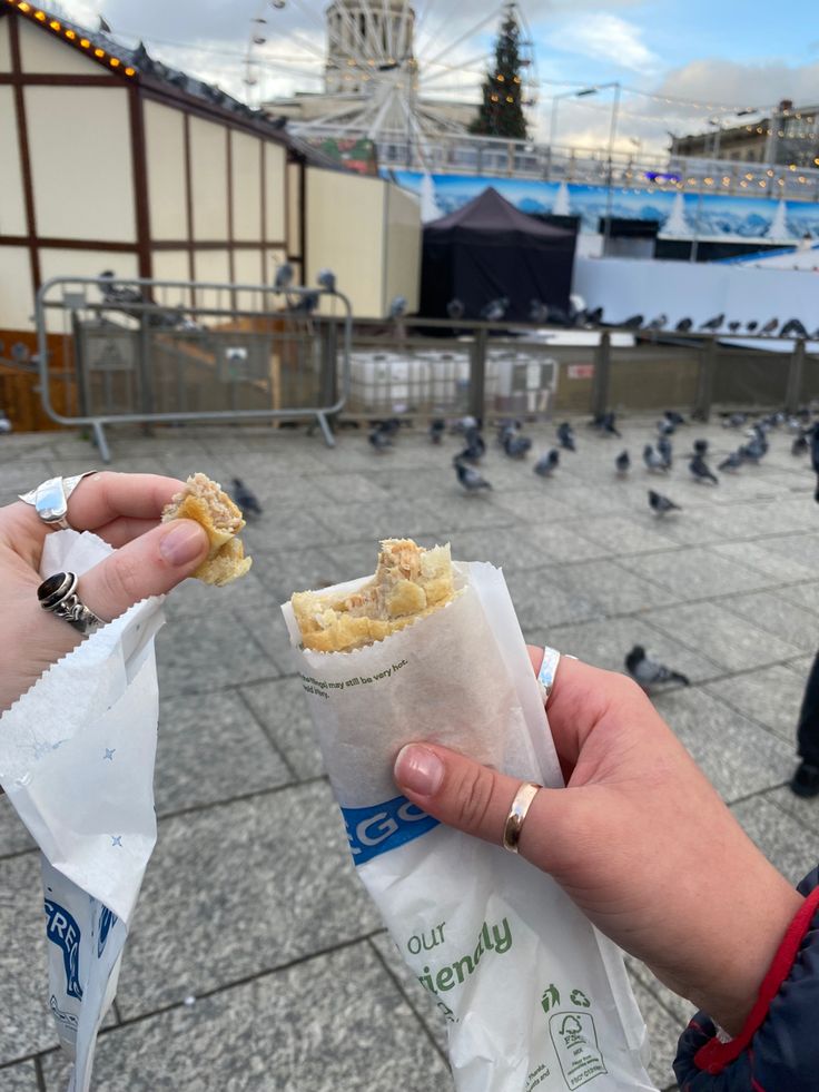 a person holding food in their hand while pigeons fly around the area behind them and on the ground