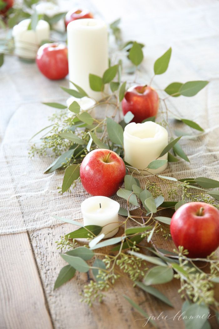 an arrangement of apples, candles and greenery on a linen tablecloth with leaves