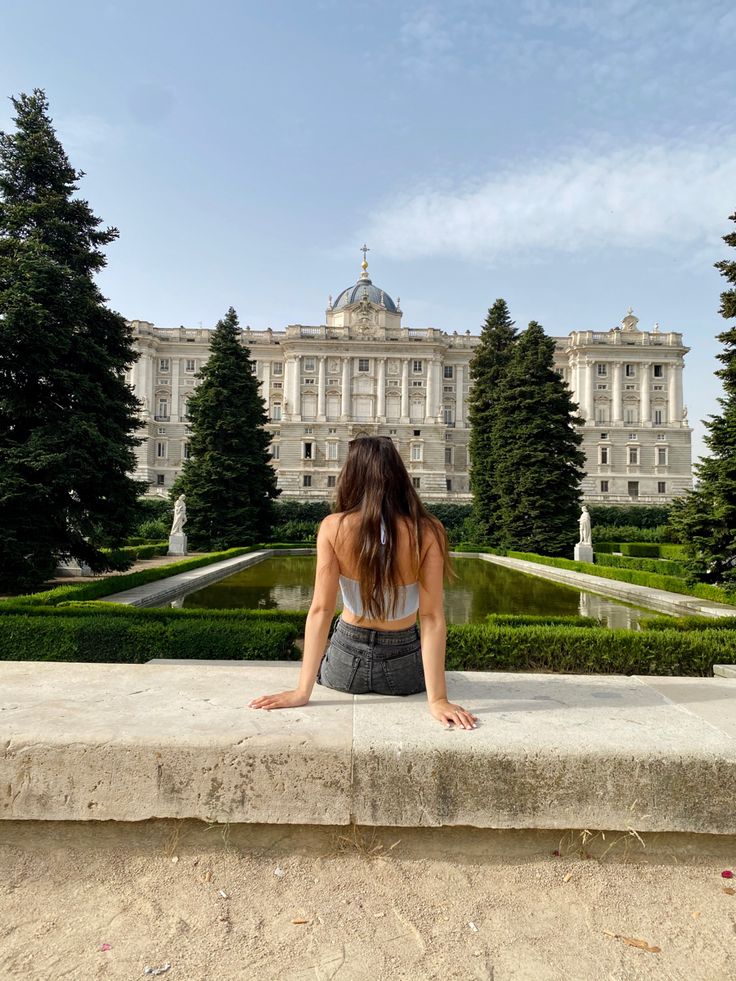 a woman sitting on a ledge in front of a large building with trees and bushes