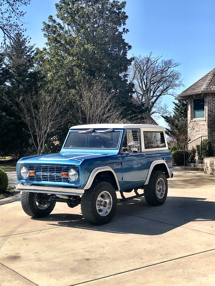 a blue jeep is parked in front of a house with trees and bushes behind it