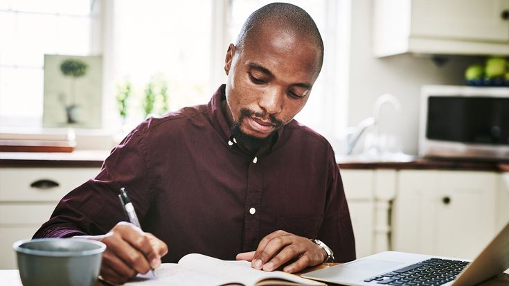 a man sitting at a kitchen table working on his laptop and doing paperwork with a cup of coffee in front of him