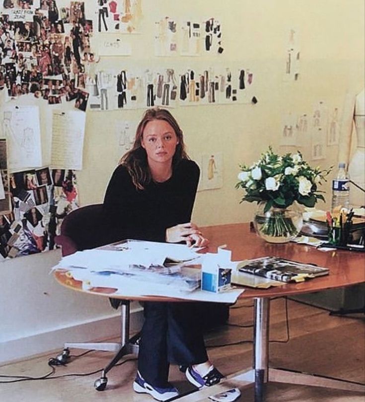 a woman sitting at a table in front of a wall covered with pictures and papers