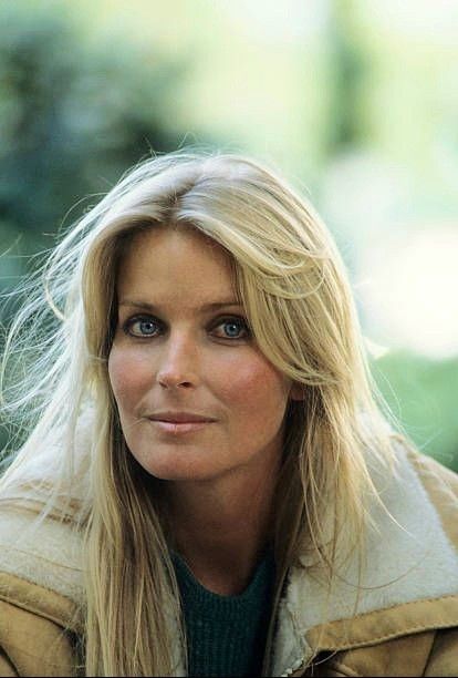 a woman with long blonde hair and blue eyes looks into the distance while wearing a brown jacket