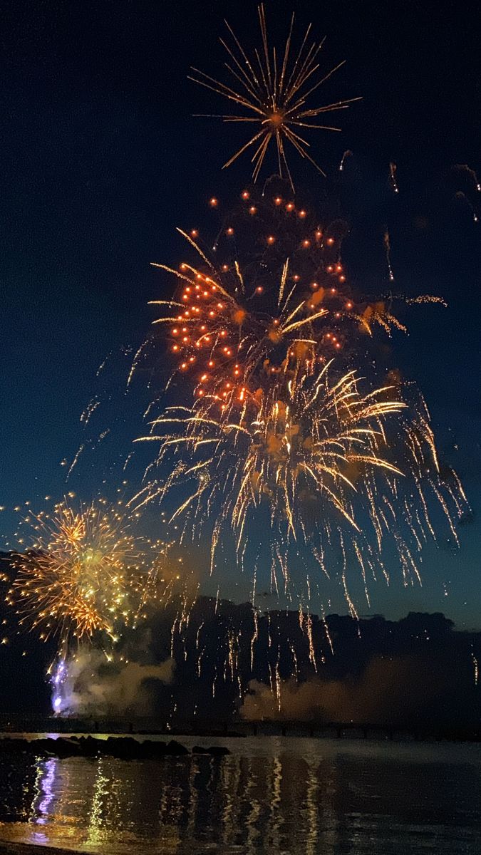 fireworks are lit up in the night sky over water and mountains as seen from a beach