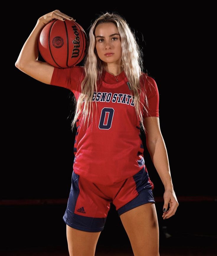 a female basketball player holding a ball in her right hand and posing for the camera