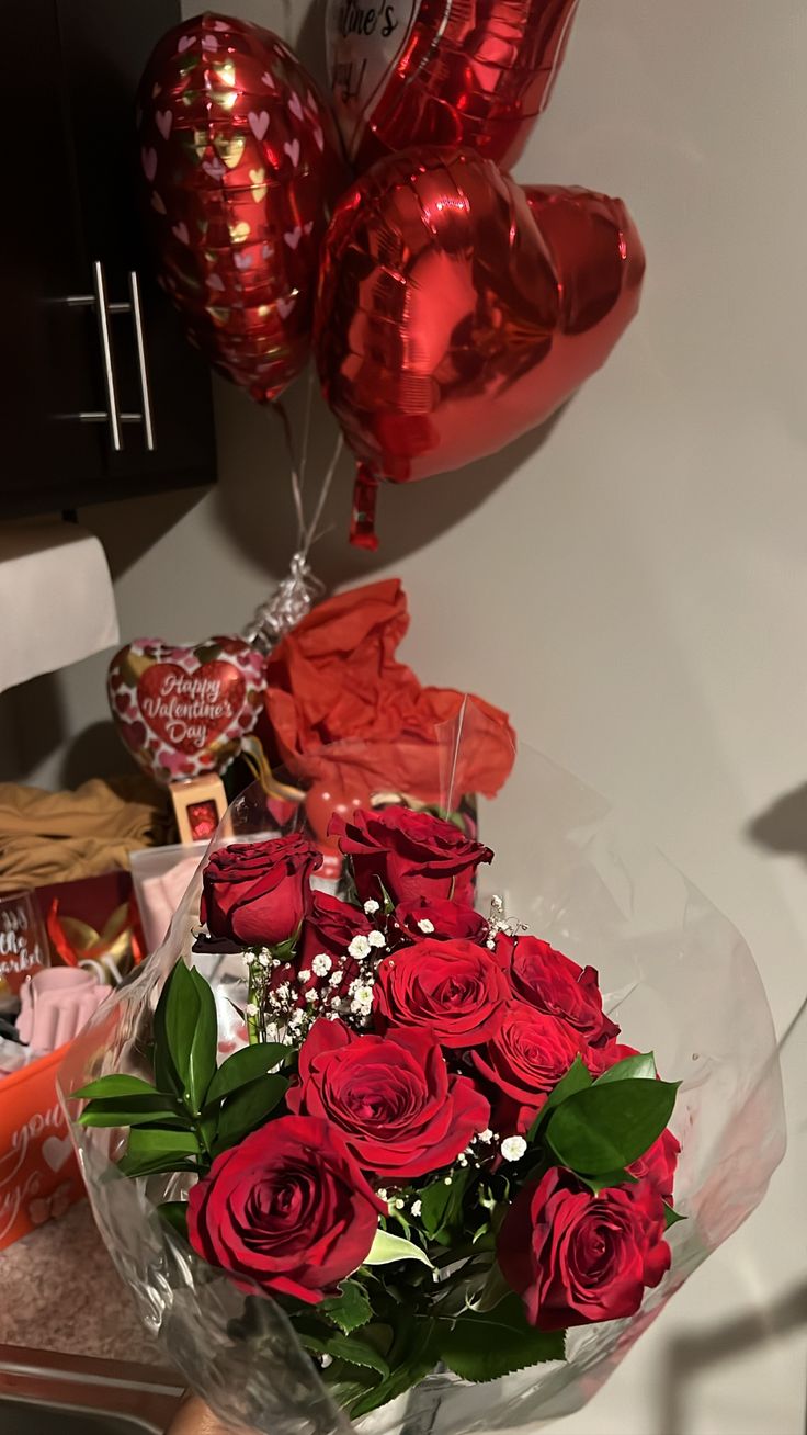 a bunch of red roses in a vase with some heart shaped balloons on the wall