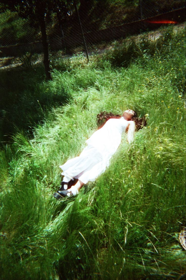 a woman laying in the grass with her head on her hands and eyes closed, wearing a white dress