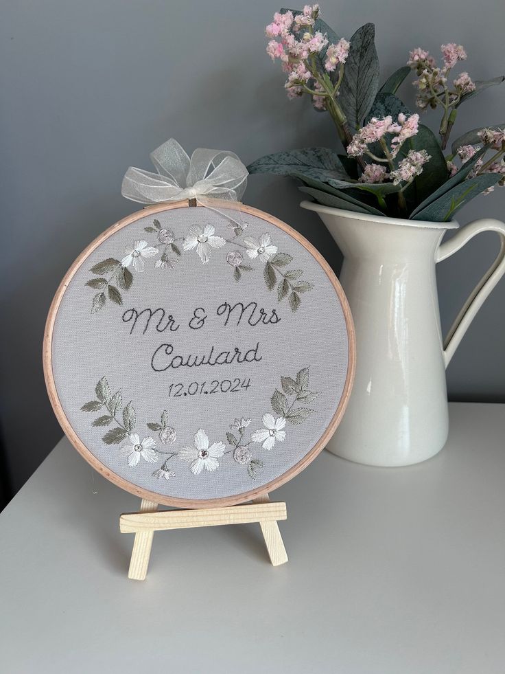 a white vase with flowers next to a cross stitched wedding ornament on a table