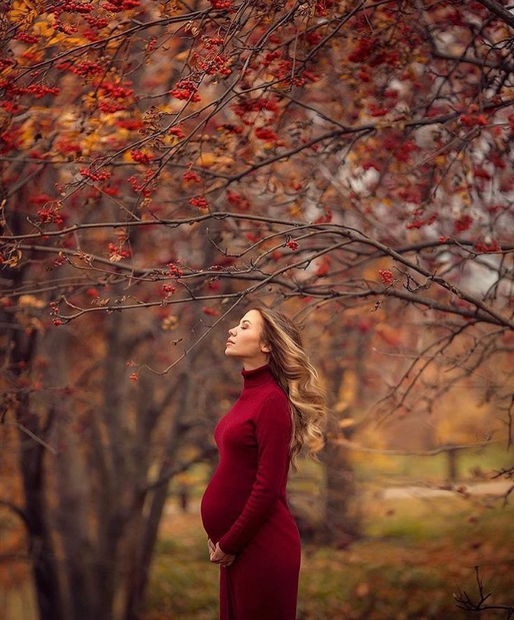 a pregnant woman in a red dress standing under a tree with fall leaves on it