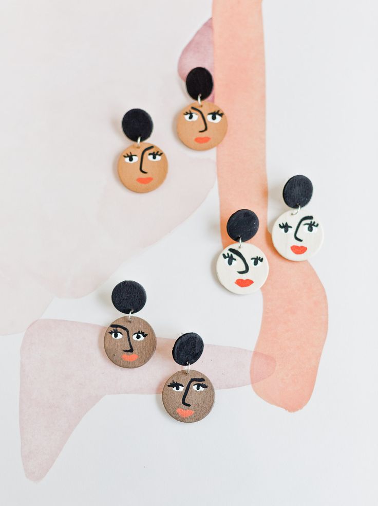 three pairs of earrings with faces hanging from the back of one ear, on top of a pink and white background