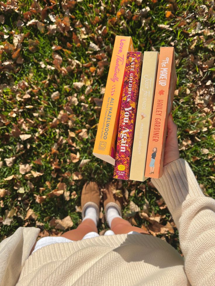 a person is holding three books in their hands while sitting on the grass with leaves around them