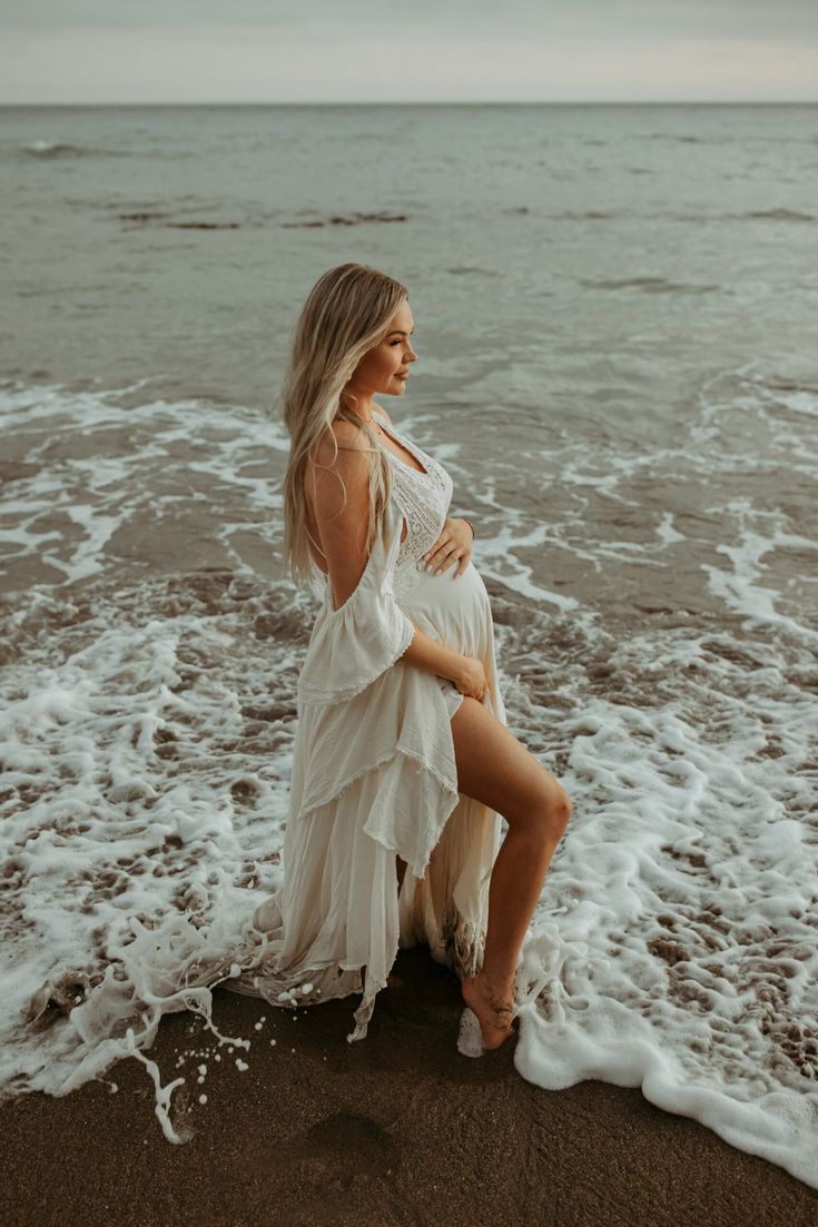 a pregnant woman sitting on top of a sandy beach next to the ocean with waves