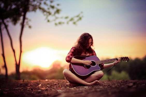 a woman sitting on the ground playing an acoustic guitar at sunset with trees in the background