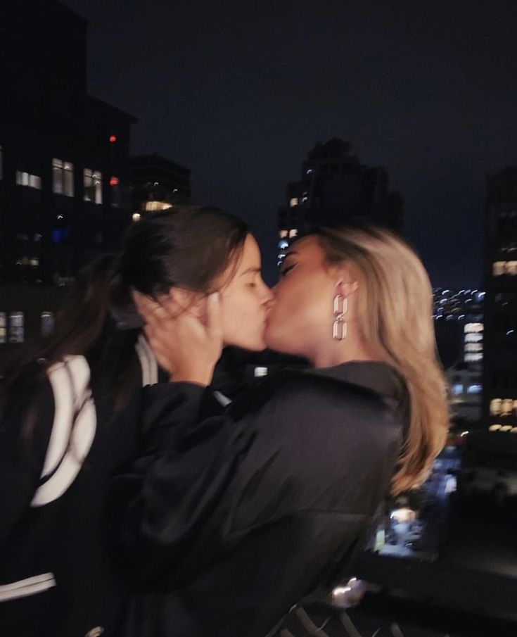 two women kissing each other in front of a cityscape at night with skyscrapers lit up behind them