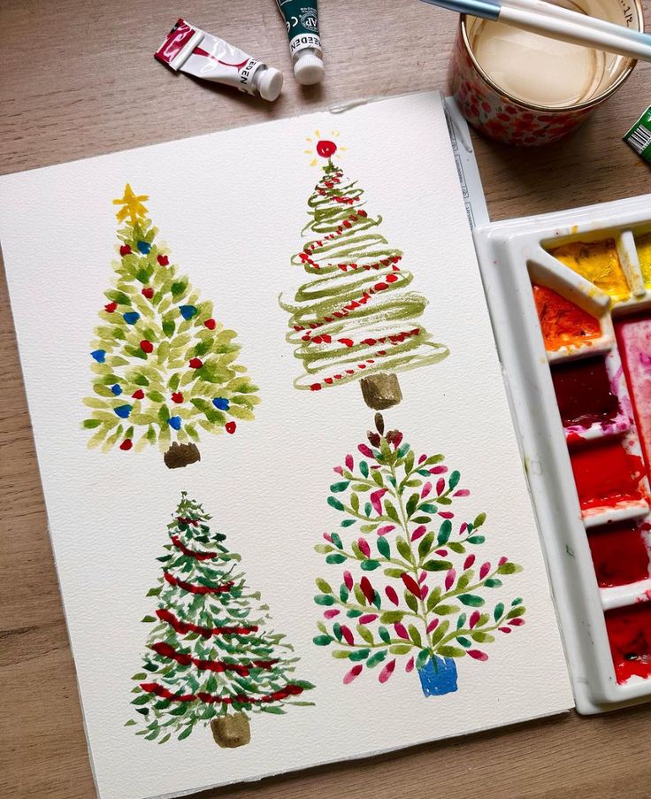 watercolor christmas trees are on the table next to paintbrushes and a cup