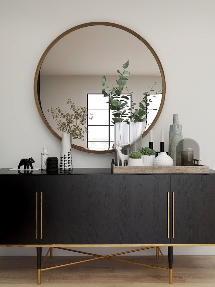 a large round mirror on the wall above a black cabinet with gold legs and handles