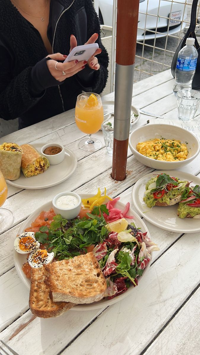 a woman is looking at her cell phone while sitting at an outdoor table full of food