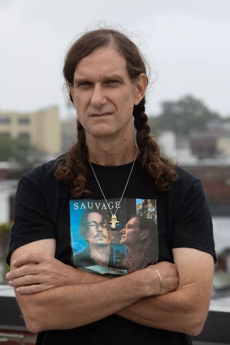 a man with long hair wearing a black t - shirt and holding his arms crossed