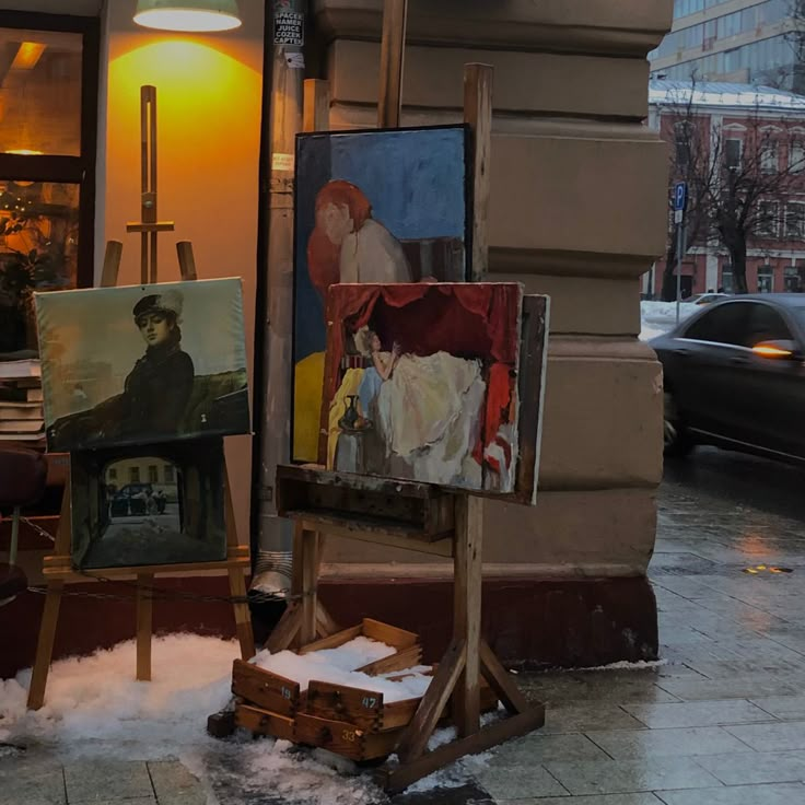 an artist's easel sitting on the sidewalk in front of a building with paintings