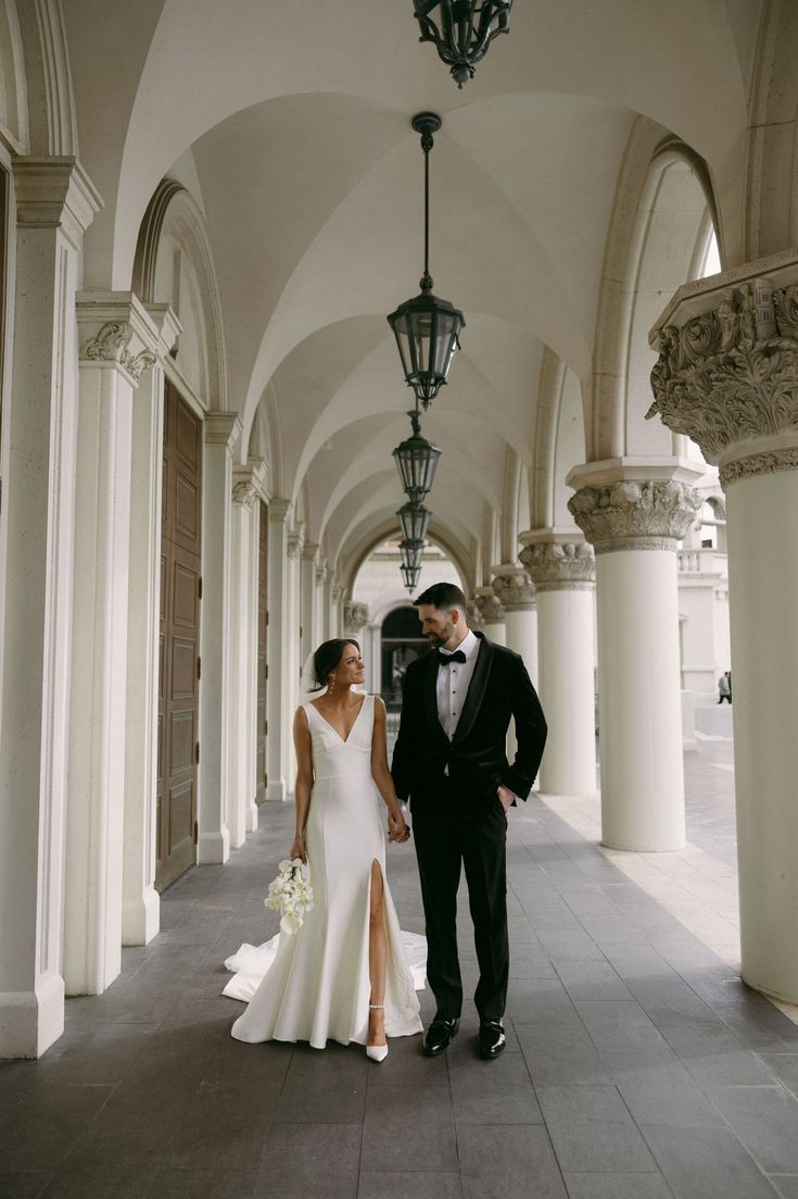 a bride and groom are standing in an arched hallway holding hands, looking at each other