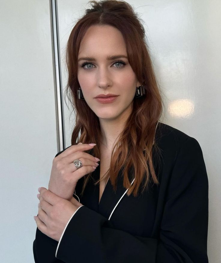 a woman with red hair wearing a black jacket and diamond ring, standing in front of a white wall