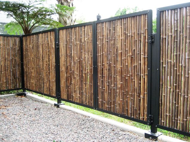 a fence made out of bamboo sticks in front of a gravel area with trees and bushes behind it