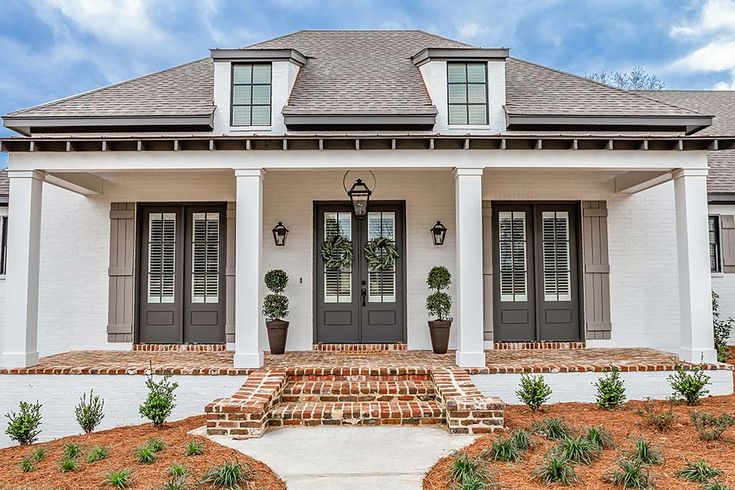 a white house with black shutters and two large doors on the front porch, surrounded by greenery