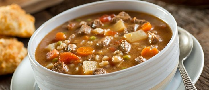 a white bowl filled with meat and vegetable soup on top of a plate next to bread