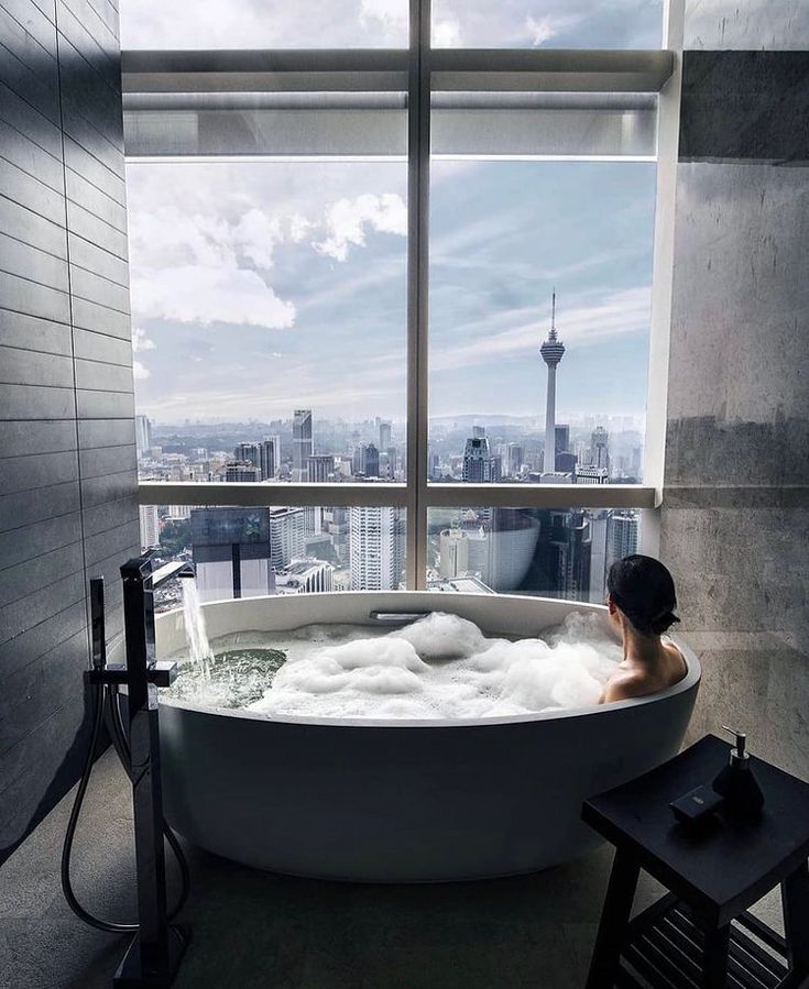 a woman sitting in a bathtub with the city skyline outside it and looking out the window