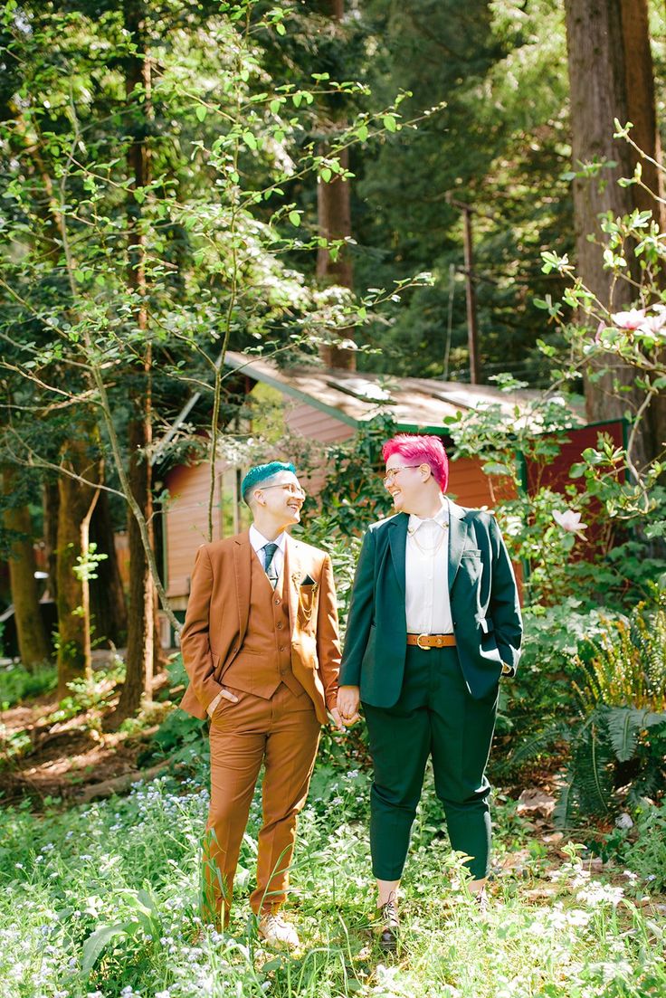 two young men standing next to each other in the grass near trees and bushes, one with pink hair