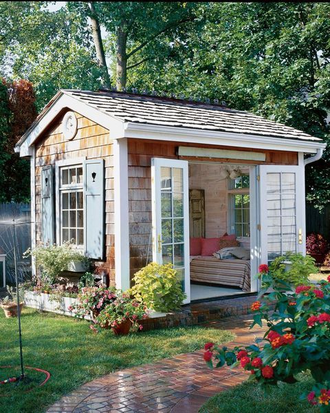 a garden shed with the doors open and flowers around it, in front of trees