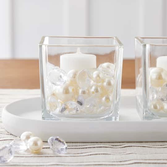 two square glass vases filled with white candles on a tray next to pearls and crystal beads