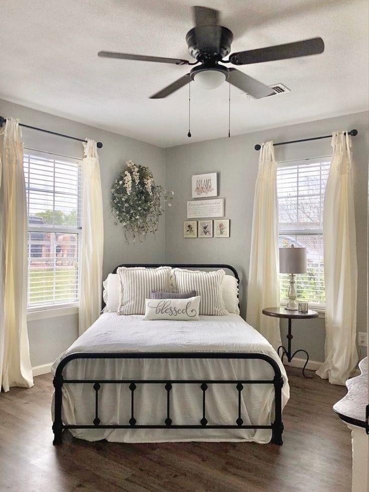 a bed sitting in a bedroom next to two windows with white drapes on them