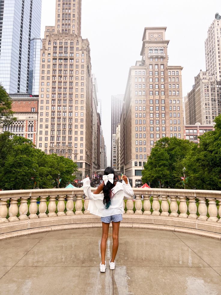 a woman standing in the middle of a city with tall buildings behind her and holding an umbrella over her head