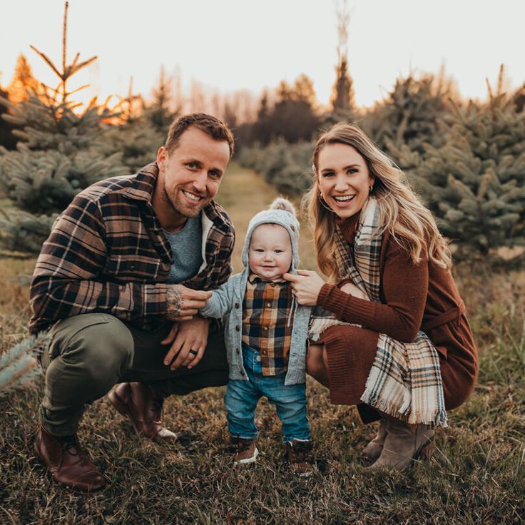 a man and woman kneeling down next to a baby in a field with christmas trees