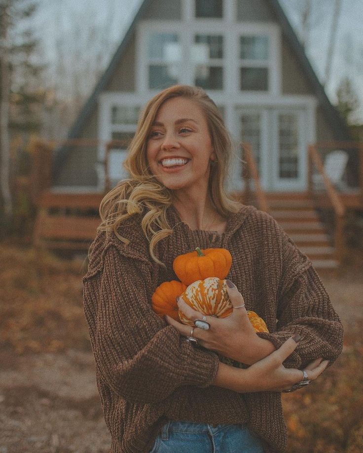 a woman is holding pumpkins in front of a house and smiling at the camera