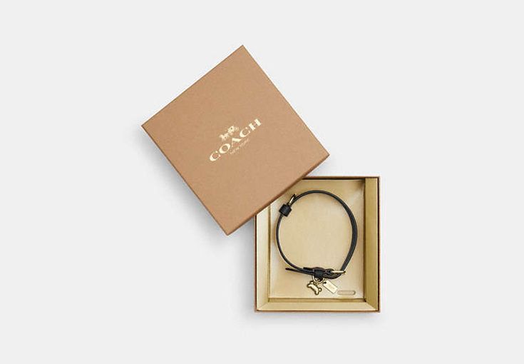 an open box with a black cord bracelet in it and a gold logo on the front