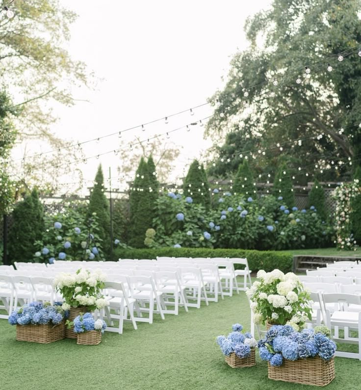 an outdoor ceremony set up with white chairs and blue flowers in baskets on the grass