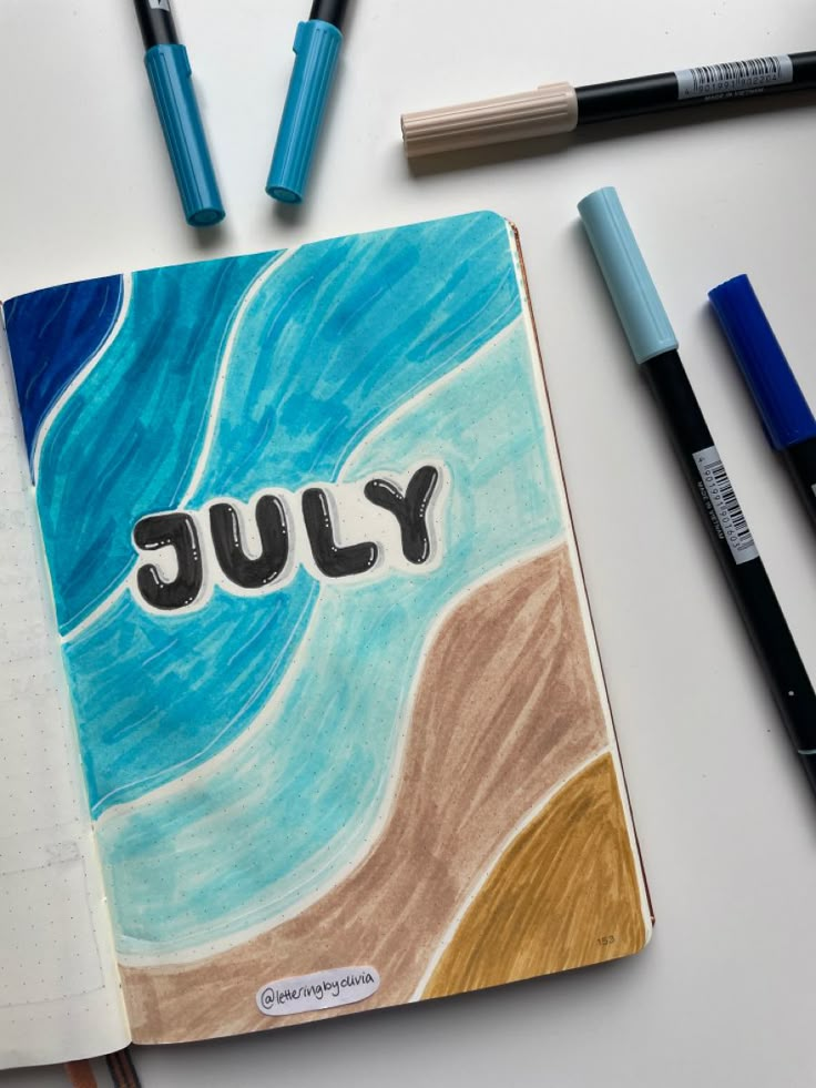 an open notebook with the word july written on it and some markers next to it