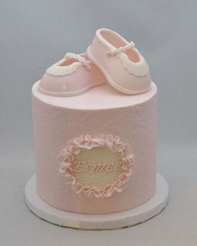 there is a pink cake with baby shoes on the top and nameplate below it