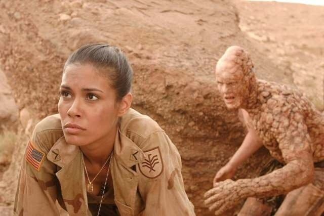 a woman in uniform sitting on the ground next to an alien creature with her hand on her chin